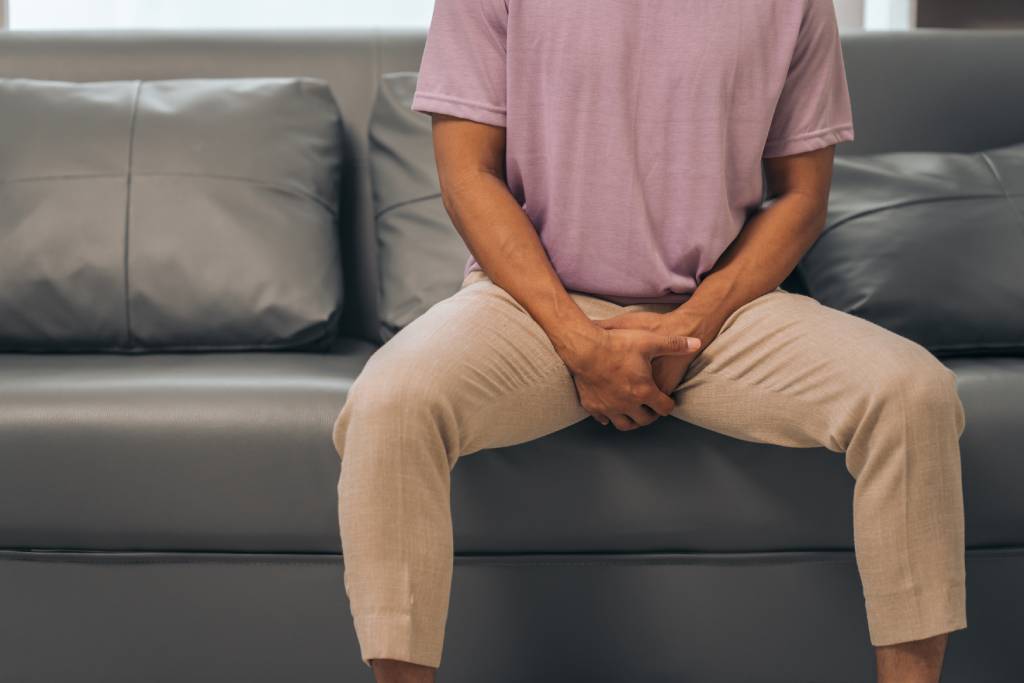 A man holds his groin in discomfort while sitting on a couch.